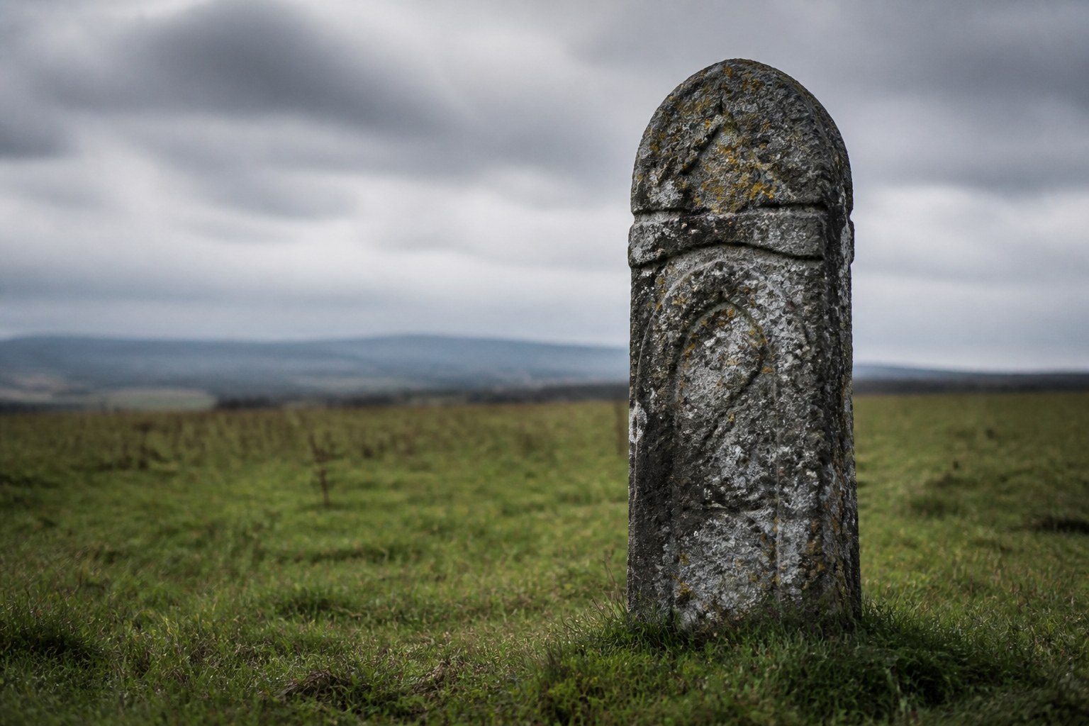 A weathered carved stone boundary marker standing alone in an open grass field under an overcast sky