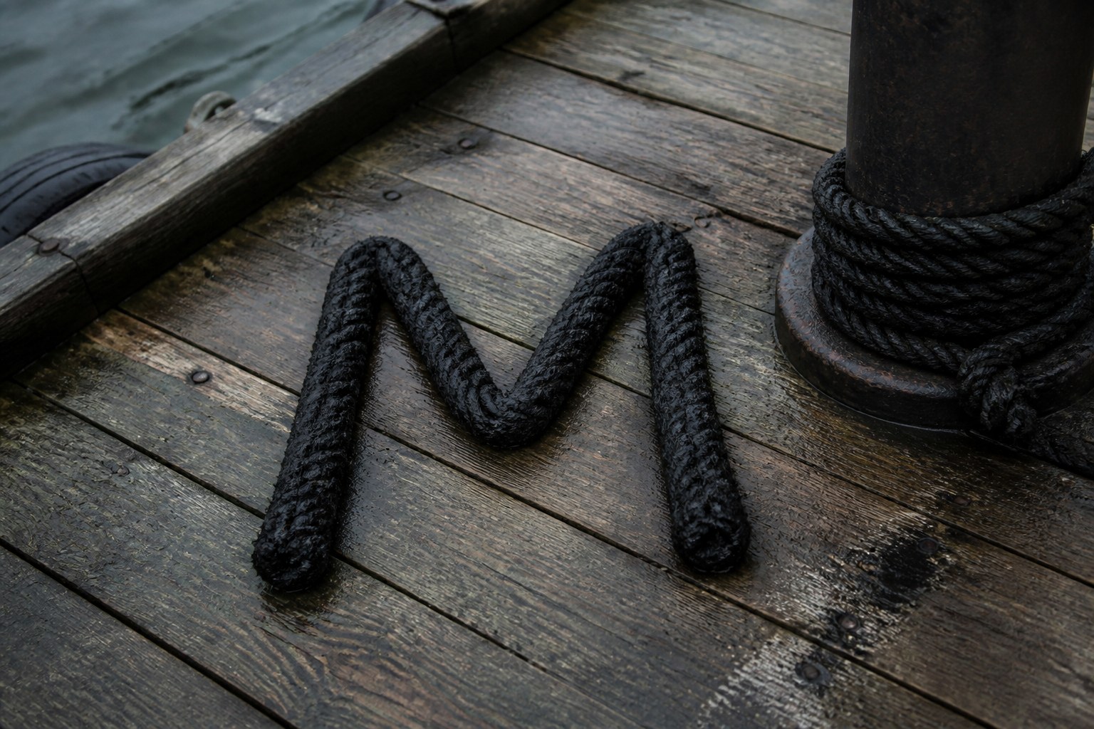 The capital letter M laid out in coiled tarred rope on weathered dock planking