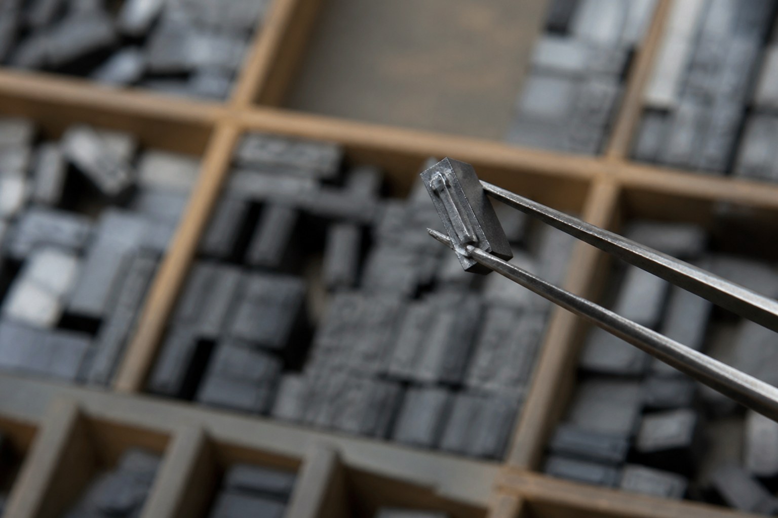A single metal type sort held in tweezers above a printer's type case