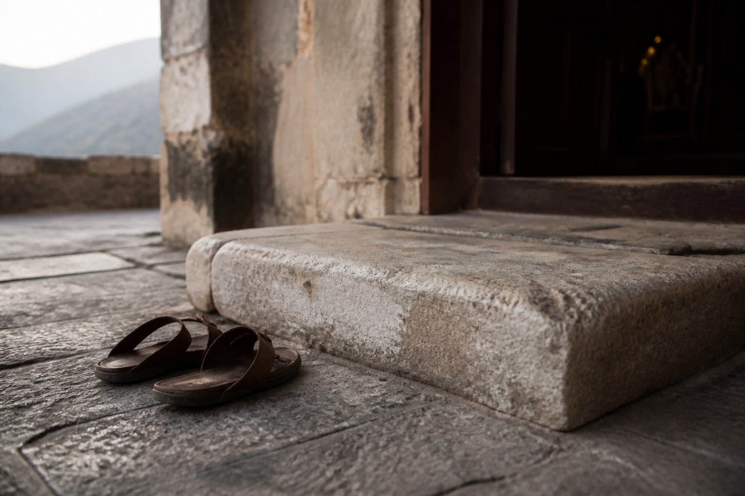 A worn stone threshold at a monastery entrance with a pair of plain sandals left beside it in morning light