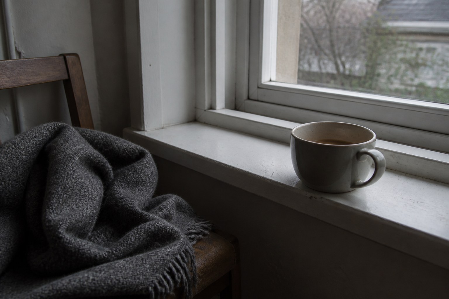 A mug of tea gone cold on a windowsill beside a wool blanket left over the back of a chair, grey late-winter daylight through the glass