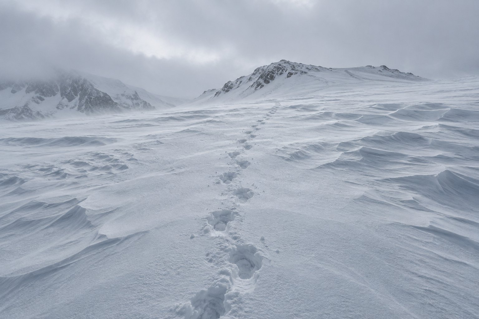 A single line of boot tracks crossing a wind-scoured snow slope toward a distant ridge under flat overcast light