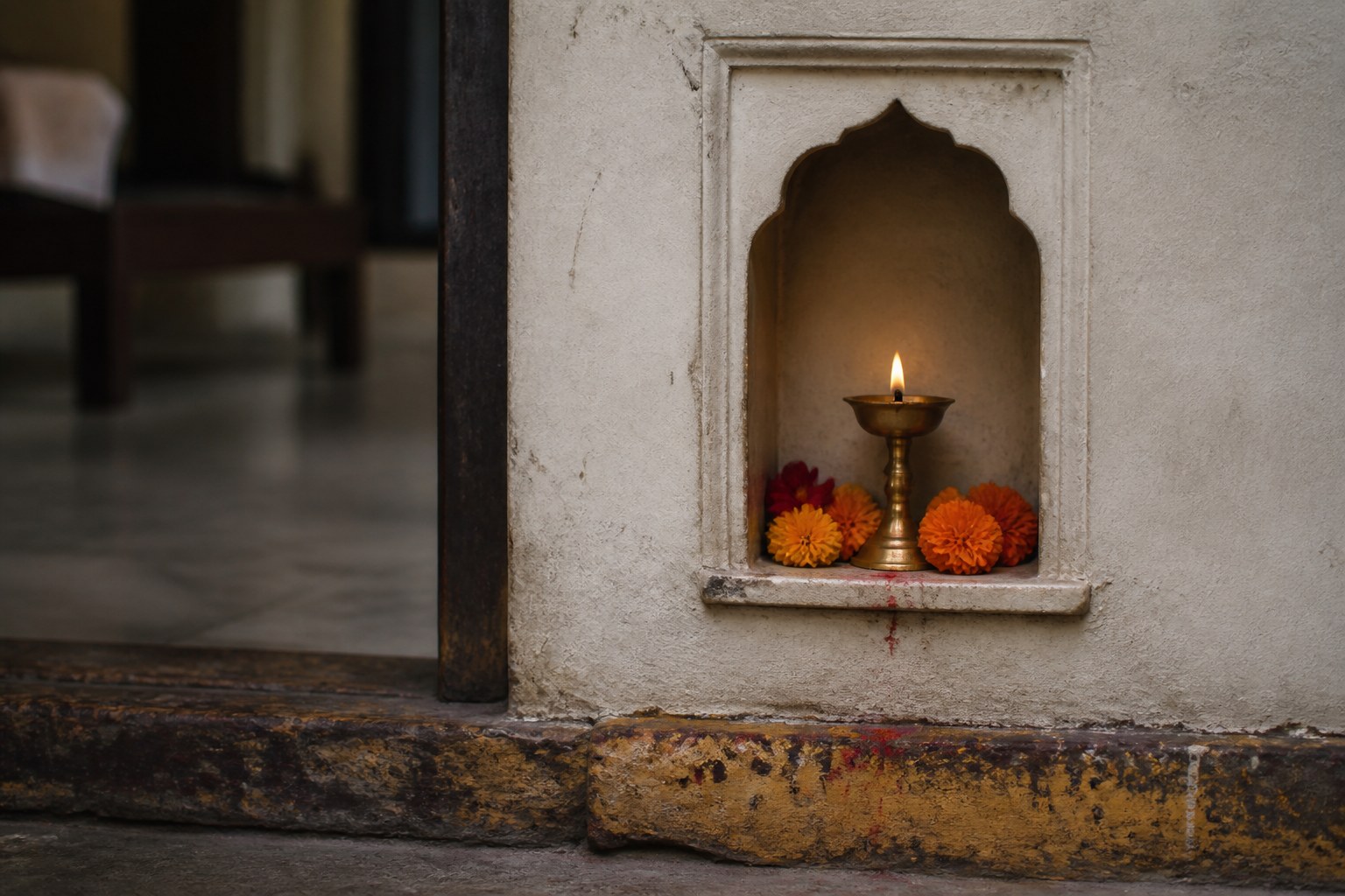 A small shrine niche set into a plastered house wall holding a lit brass oil lamp and fresh marigolds, above a worn painted threshold