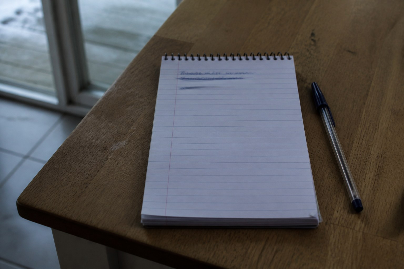 A spiral notepad on a kitchen table in cold morning light, the first two lines crossed through in ballpoint and the rest of the page still empty, a pen lying beside it
