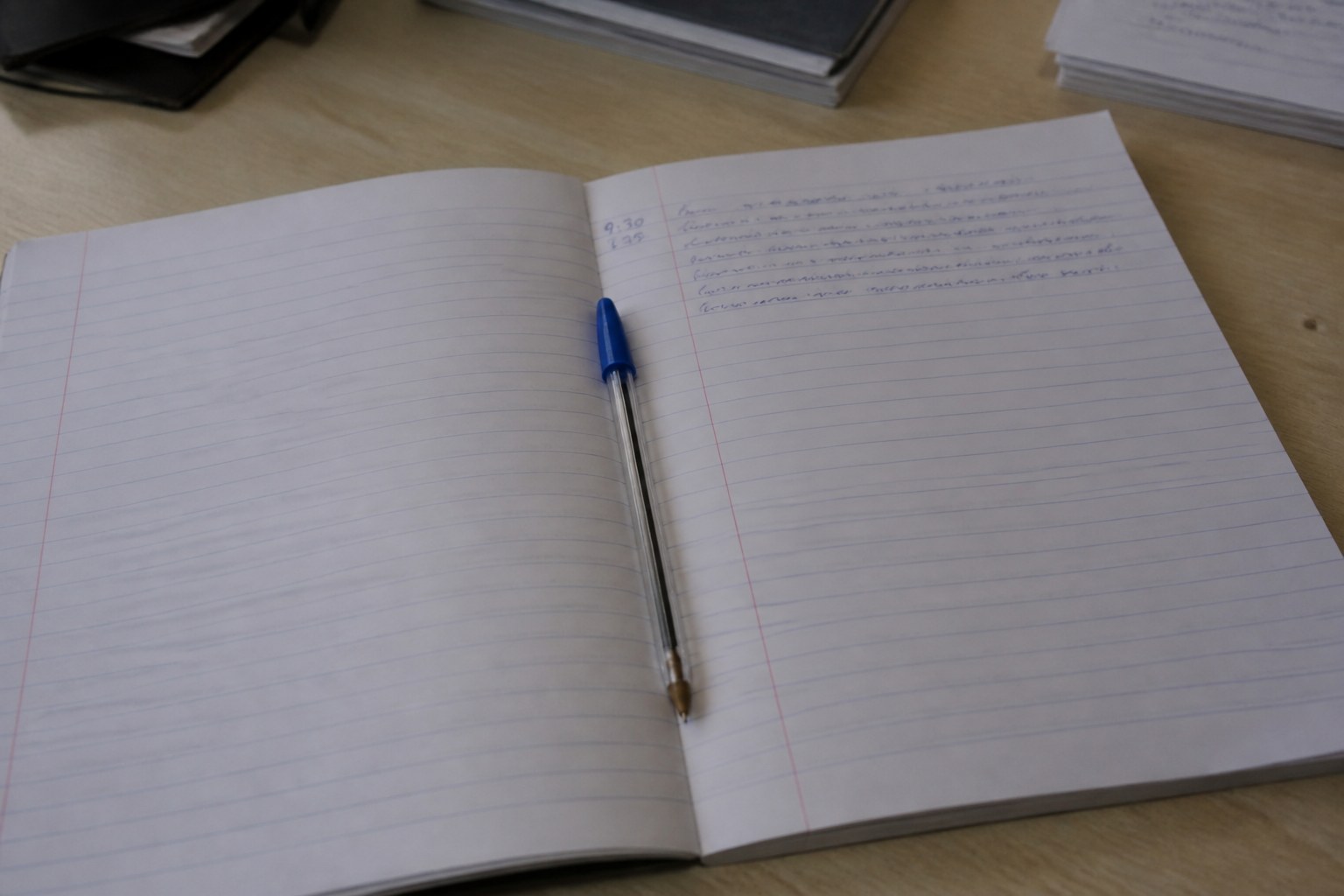 A ruled notebook open on a desk in flat daylight, a few short handwritten lines with a time noted in the margin, a biro resting across the page