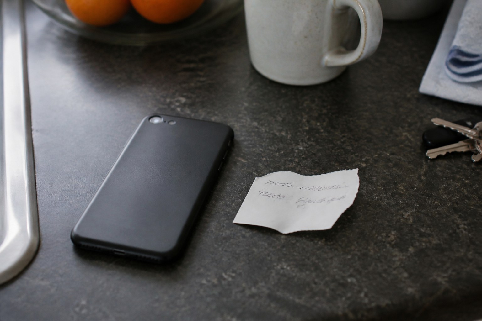 A mobile phone face down on a kitchen worktop beside a torn scrap of paper with a few words written on it, the writing not legible, keys and a mug at the edge of frame