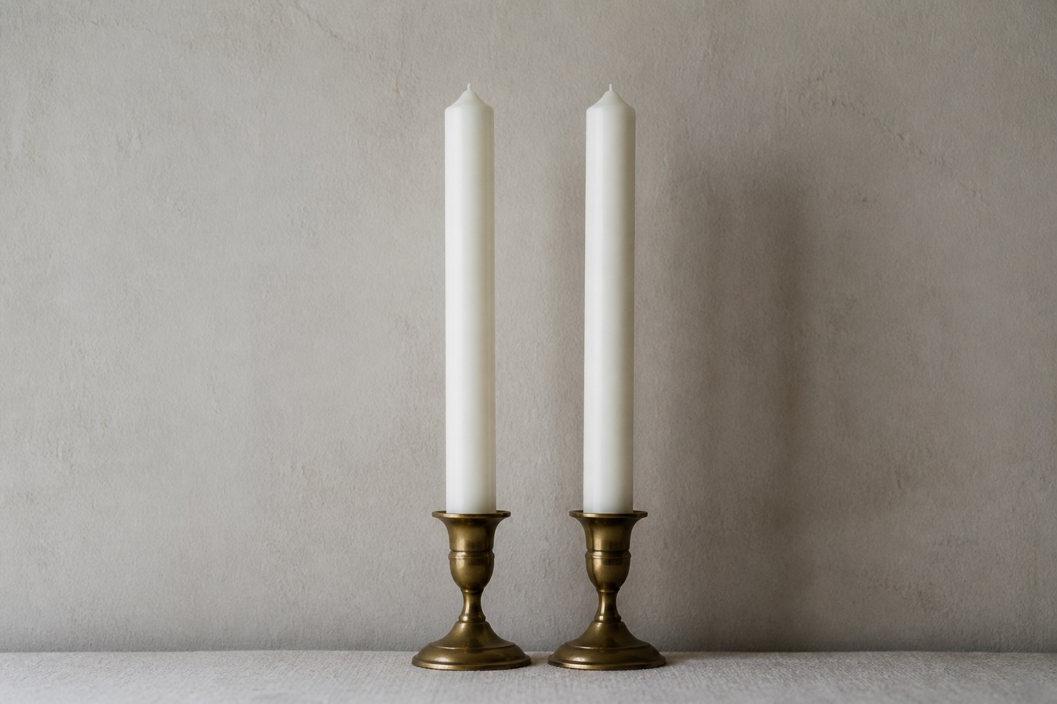 Two identical tall white church candles standing side by side, unlit, against a plain wall