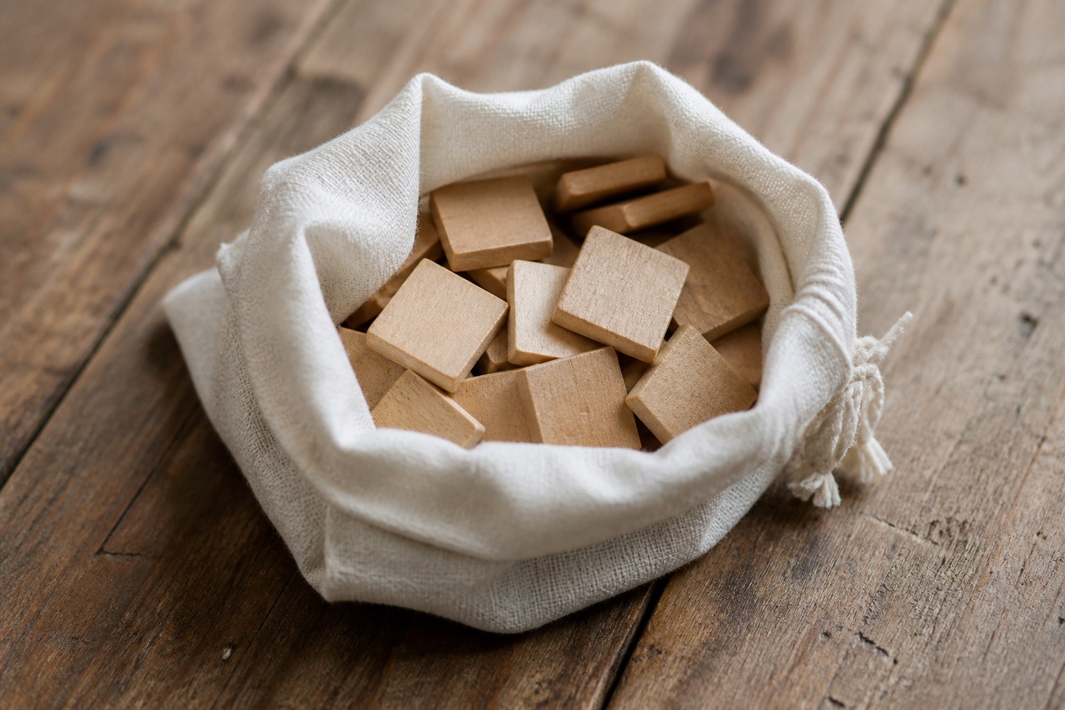 Wooden letter tiles lying face down in an opened drawstring bag on a table, their blank backs upward