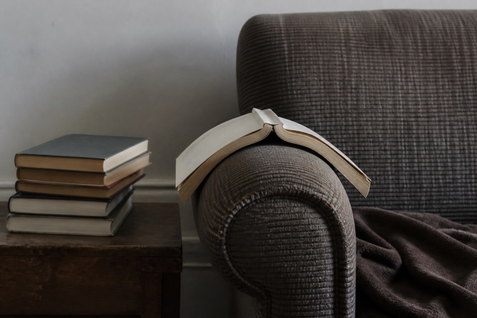 A single book left open face down on a chair arm with an unopened pile beside it