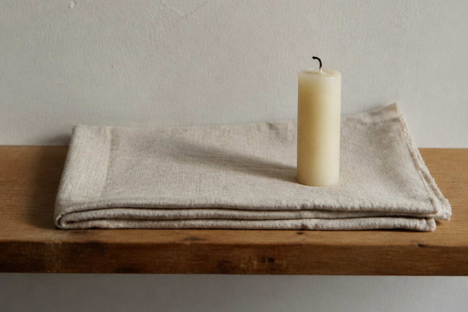 A narrow wooden shelf against a pale plaster wall, bare except for a folded length of undyed linen and one unlit cream candle with a blackened wick