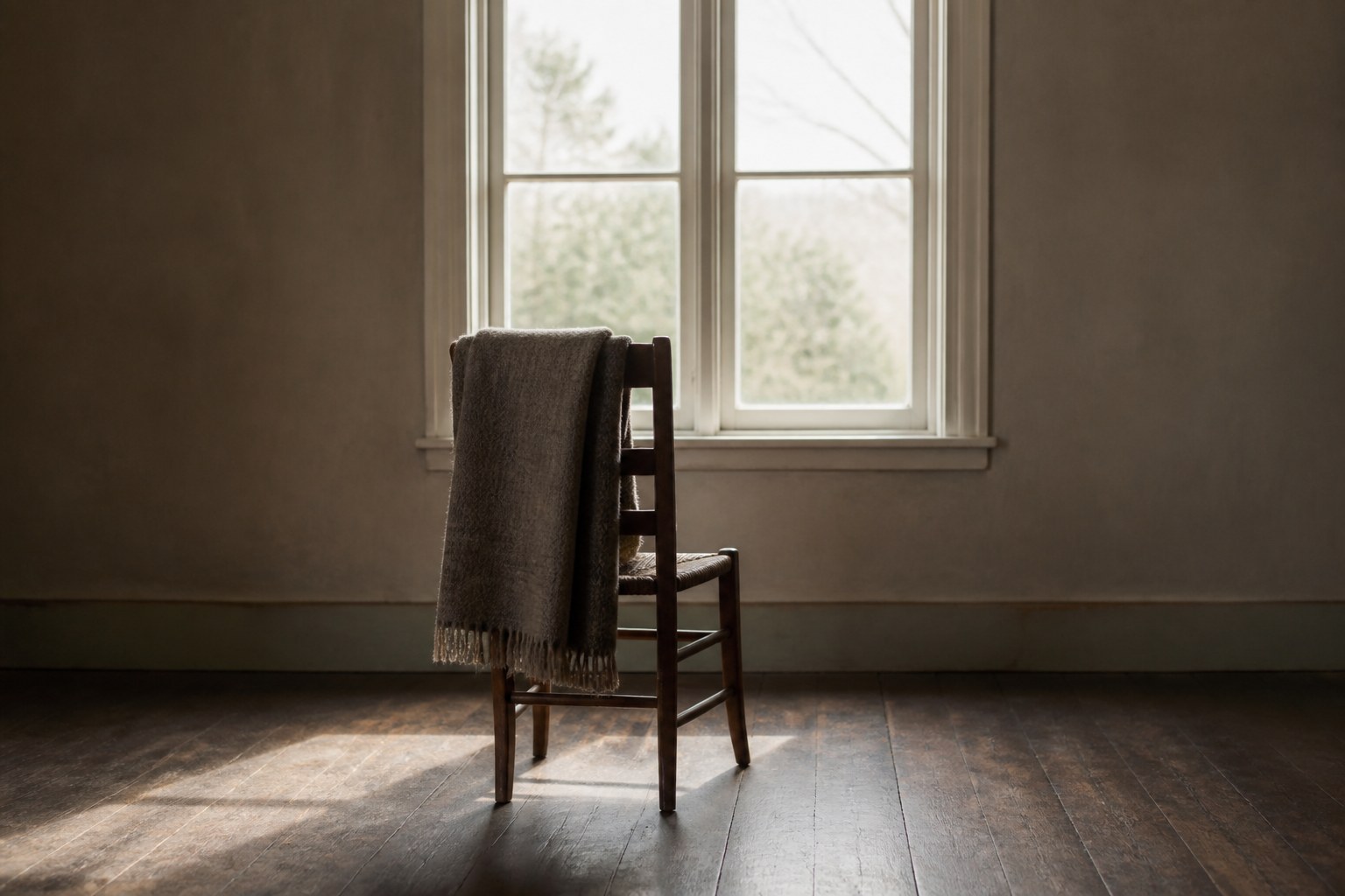 A plain upright wooden chair facing a bright window in an empty room, a folded wool blanket over its back