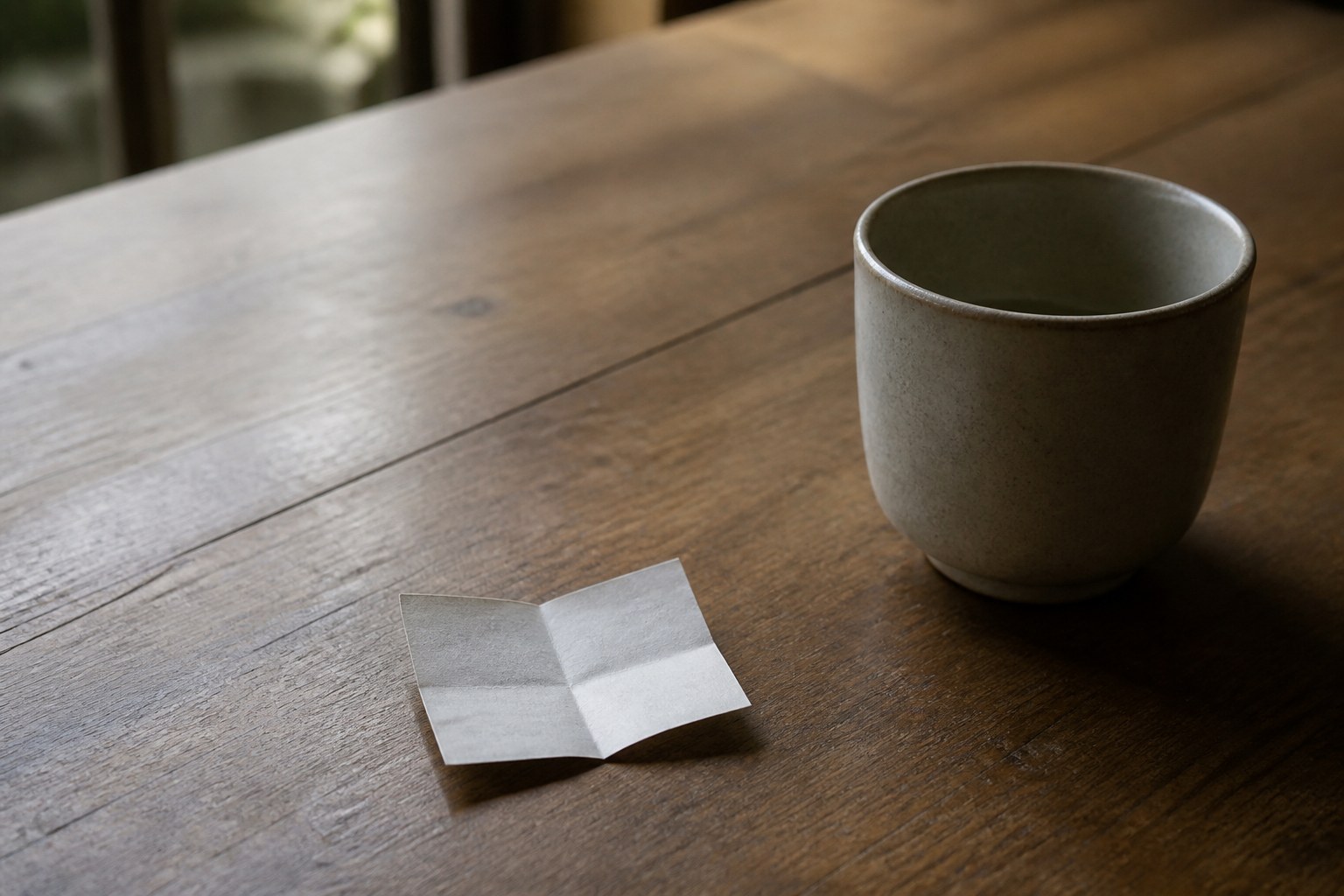 A small folded paper slip on a plain wooden table beside an empty cup, the writing inside not visible