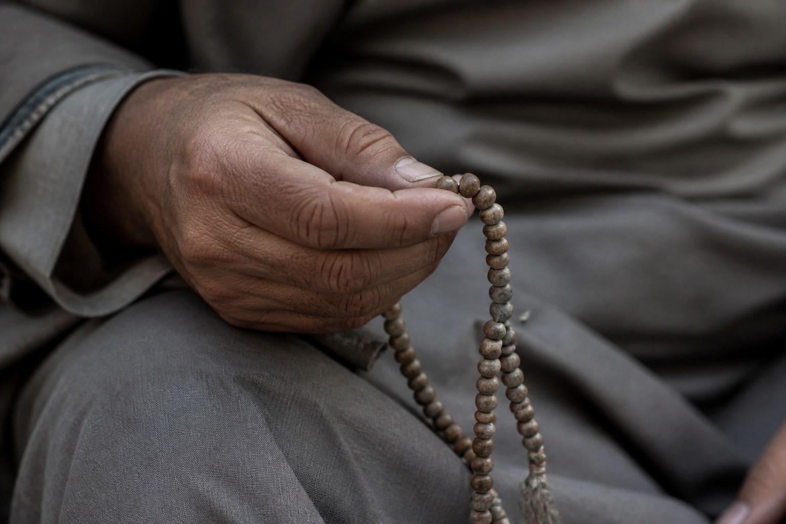 A seated person's hand holding a strand of prayer beads with the thumb resting on a single bead, the rest of the strand hanging loose